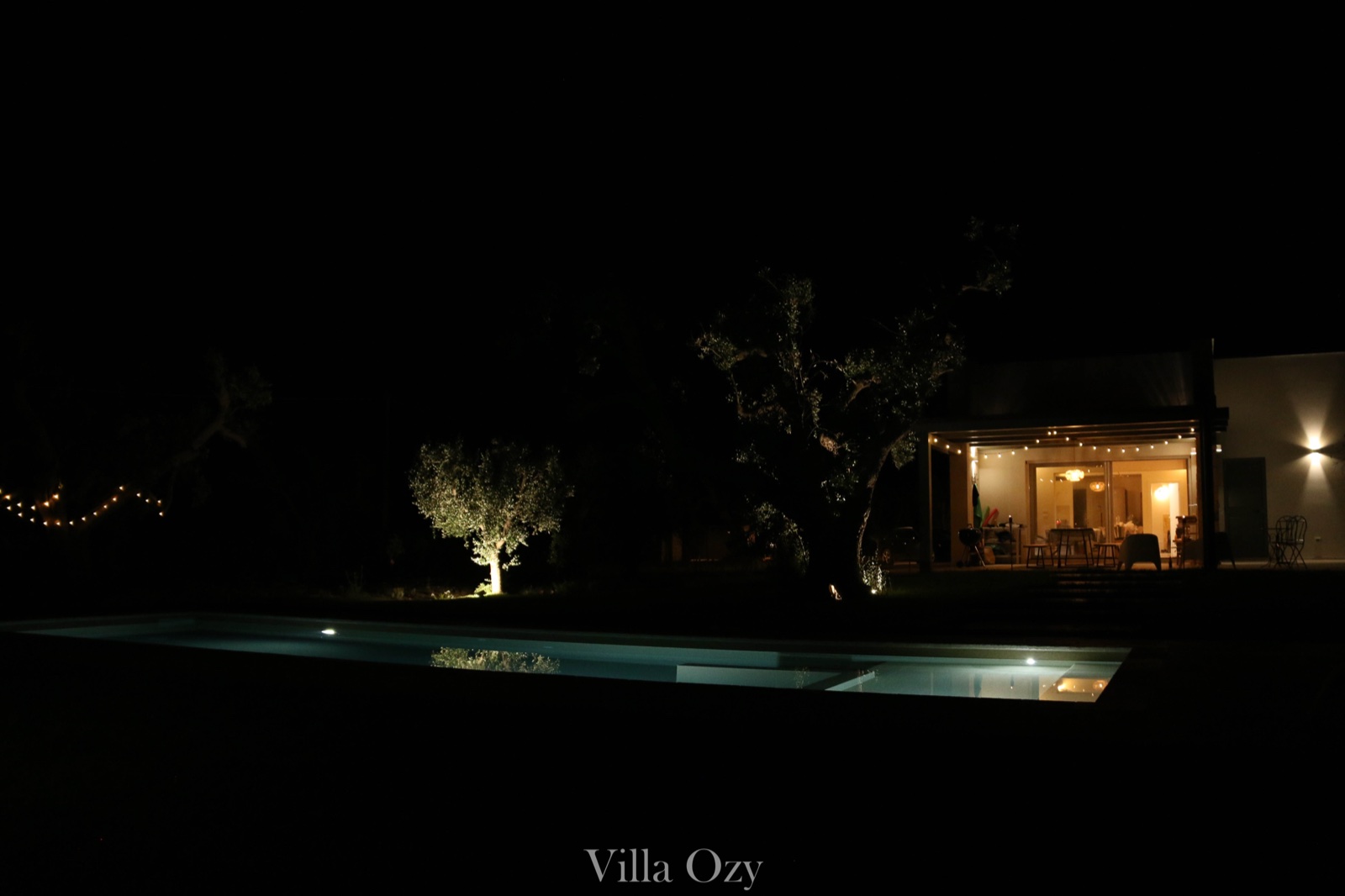 Villa Ozy at night, pool and olive trees softly lit beside the lit interior